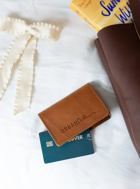 An Urban Southern Bifold Card Wallet made of brown full-grain leather rests beside a blue Discover card, with a brown bag containing yellow books and a cream ribbon set on a white surface.