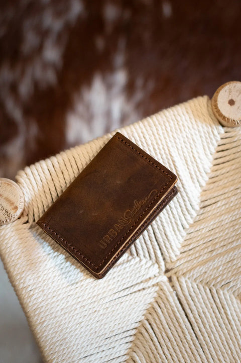 The Urban Southern Bifold Card Wallet, crafted from full-grain leather, is displayed on a woven cream seat with a blurred brown and white background.