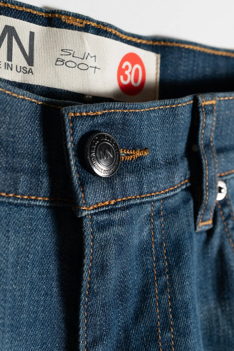 Close-up of The Boot Cut Medium Wash Denim by Marc Nelson, featuring a branded metal button, orange stitching, and a white SUM BOOT label for boot cut jeans—size 30 highlighted in red and MADE IN USA partially visible.
