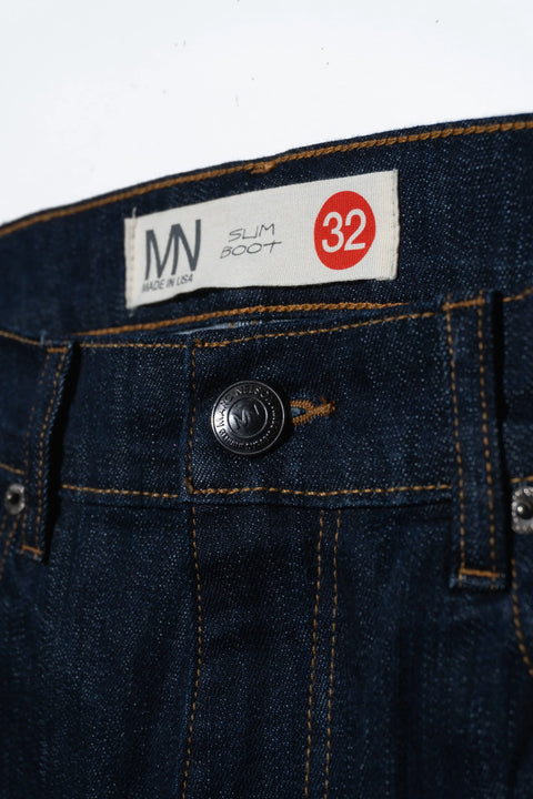 Close-up of the waistband on Marc Nelsons The Boot Cut Dark Wash Denim, featuring a black button, orange stitching, and a label reading MN Made in USA Slim Boot 32 with the size highlighted in red.