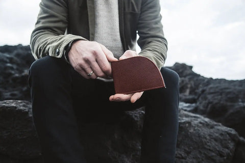 Outdoors on dark rocks beneath a cloudy sky, a person in a green jacket, light sweater, and black pants holds the Rogue Industries Football Leather Front Pocket Wallet crafted from textured Horween leather.