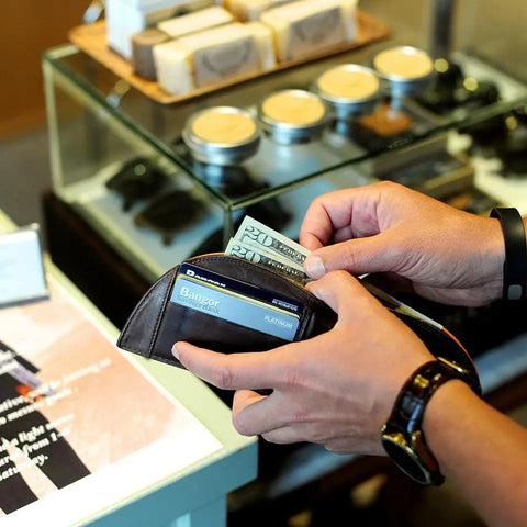 Someone holds an open Rogue Industries Football Leather Front Pocket Wallet with a Bangor ID and $20 bill, ready to pay at a counter where soaps and jars are displayed in the background.