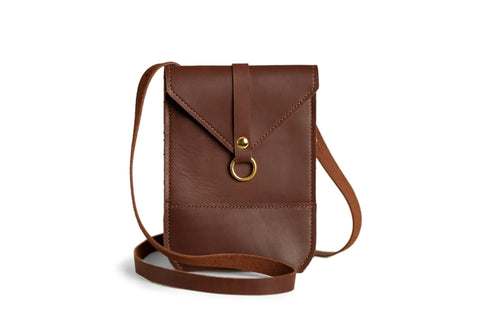 The Urban Southern Postal Crossbody features a minimalist leather design with a flap closure, gold ring detail, and long strap, shown standing upright on a white background.