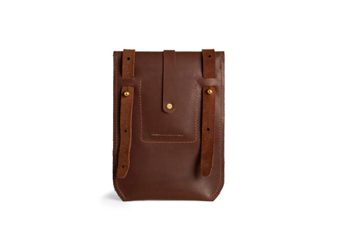 The Postal Crossbody by Urban Southern is a brown leather bag with two straps and brass button details, shown against a white background. This minimalist design combines timeless style with everyday practicality.