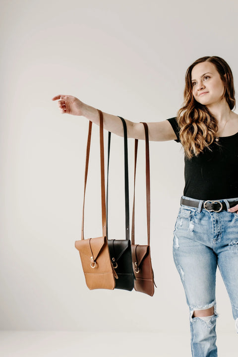 A woman in a black shirt and ripped jeans holds out her arm, showing three Urban Southern Postal Crossbody bags in brown and black, each minimalist bag elegantly hanging from her arm against a plain light background.