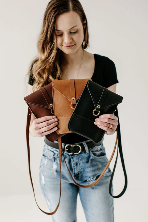 A woman in a black top and ripped jeans holds three Urban Southern Postal Crossbody bags—in brown, tan, and black—displayed in front of her against a plain white background.