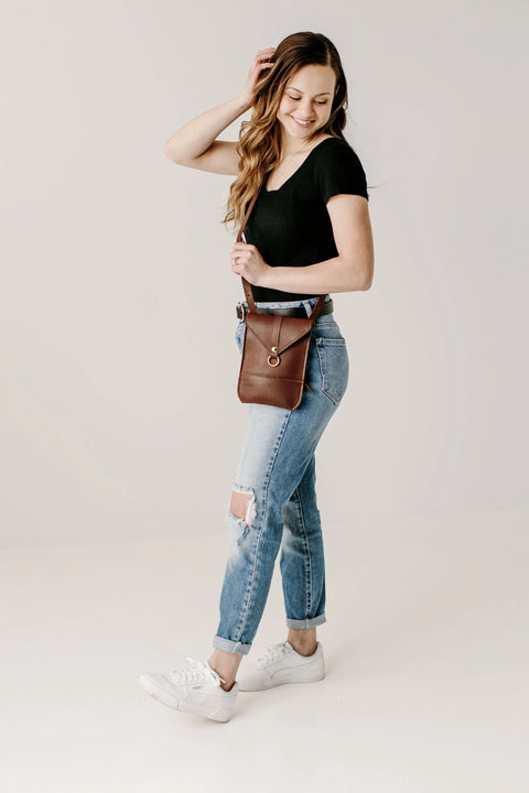 A woman smiles in a black t-shirt, ripped blue jeans, white sneakers, and the Urban Southern Postal Crossbody. With long hair and one hand touching her head, she poses against a plain white background.