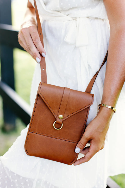 A woman in a white dress holds the Urban Southern Postal Crossbody, a small minimalist bag with a gold ring clasp. She wears a gold bracelet and light nail polish. The blurred outdoor background highlights the chic style.