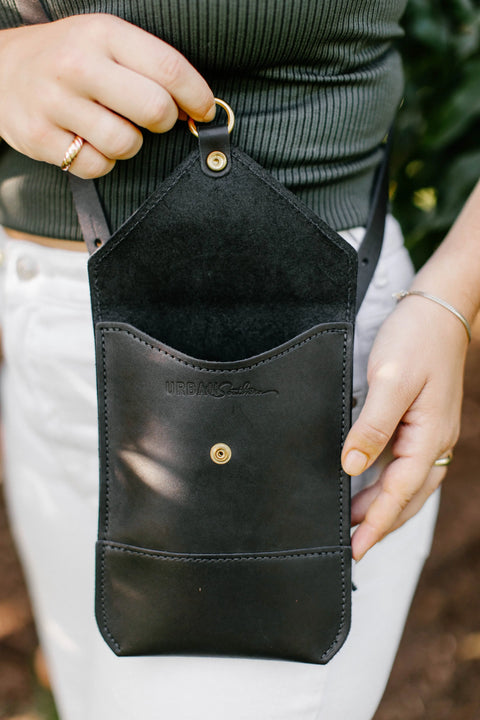 A person holds an open black Postal Crossbody bag by Urban Southern with a gold snap button, wearing a ribbed green top, white pants, and gold jewelry.