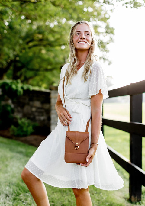 A smiling woman in a white dress stands outdoors near a wooden fence and green trees, holding the Urban Southern Postal Crossbody. Sunlight brightens the scene, creating a cheerful atmosphere.