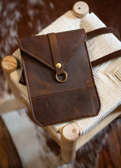The Urban Southern Postal Crossbody is an American-made brown leather bag with a brass button and ring clasp, shown resting on a woven cream stool with wooden legs—a perfect blend of style and craftsmanship.