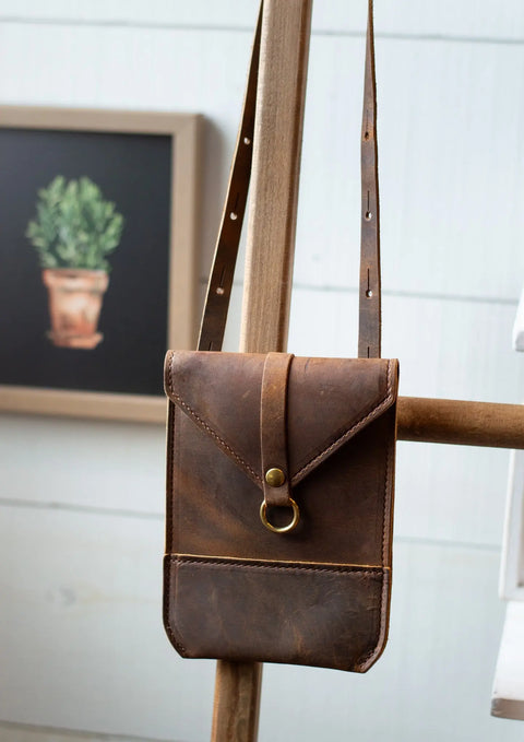 The Urban Southern Postal Crossbody, a minimalist leather bag with a brass ring clasp, hangs on a wooden easel. Behind it, a framed picture of a potted plant rests against a white wall.