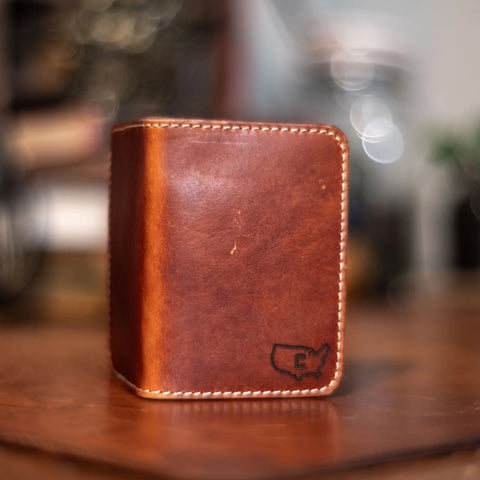 The Slim Leather Wallet – Crafted Signature 6 Pocket by KC Laser Co stands upright on a wooden surface, featuring white stitching and a USA outline with a C engraved. This American-made wallet is handcrafted; background appears blurred.