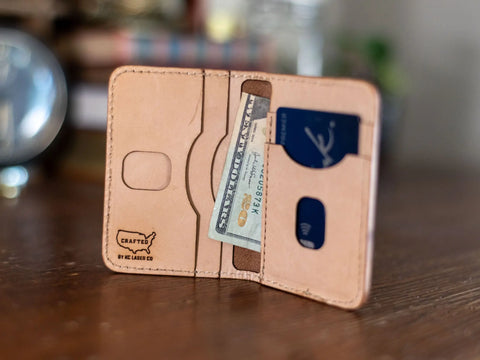 A handcrafted Slim Leather Wallet – Crafted Signature 6 Pocket by KC Laser Co rests open on a wooden surface, revealing a $100 bill and credit cards. Visible stitching and the stamped KC Laser Co logo highlight its USA-made quality.
