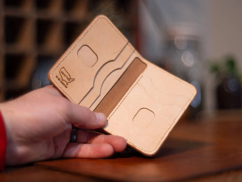A hand holds an open, empty Slim Leather Wallet – Crafted Signature 6 Pocket by KC Laser Co, made in the USA, featuring card slots and stitched edges in light tan leather against a blurred indoor background.