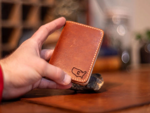 A hand holds the KC Laser Co Slim Leather Wallet – Crafted Signature 6 Pocket, made in the USA with brown leather, visible stitching, and an embossed outline of the US with a C on the bottom right. The background is softly blurred.
