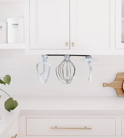 The Chatelet Manufacturing Slim Profile Mixer Organizer Mount neatly stores three Kitchen Aid mixer accessories under white cabinets, saving space in a bright kitchen. A plant and cutting board sit on the white countertop. Made in the USA.
