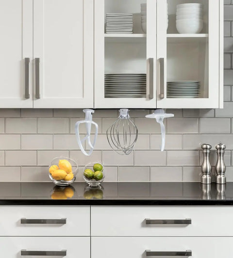 The Chatelet Manufacturing Slim Profile Mixer Organizer Mount, compatible with all Kitchen Aid mixer accessories, neatly stores three attachments under cabinets above a black countertop—a space-saving solution with bowls of citrus and salt and pepper mills below.