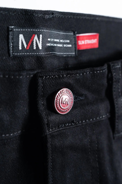 Close-up of The Nelson Black Denim waistband by Marc Nelson, made from stretch denim, with a red and silver branded button and a black MN by Marc Nelson Slim Straight label.