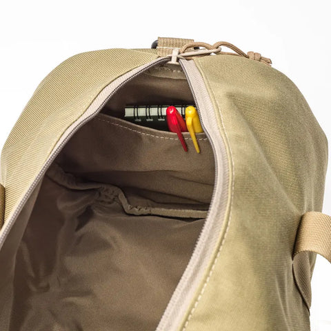 The MIS Training Drum Bag Small in Coyote Tan, made in the USA from water-resistant Cordura nylon, is unzipped to reveal an interior pocket with a small notebook and three pens—two red and one yellow. The main compartment appears empty.