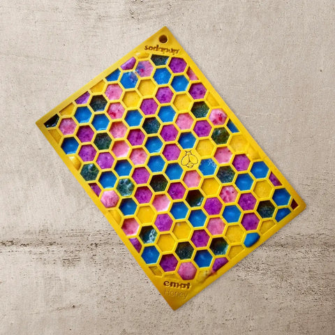 The Small Pink Flower Power & Small Yellow Honeycomb EMat Lick Mat Bundle by SodaPup features a yellow honeycomb-shaped tray with hexagonal cells filled with blue, pink, and white swirls—perfect as a dog enrichment toy.