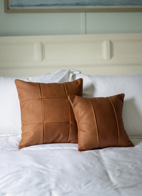 Two brown Urban Southern Small Leather Pillows—one with a stitched grid pattern, the other with vertical stitching—are arranged on a white bed featuring matching white pillows and a white headboard in the background.
