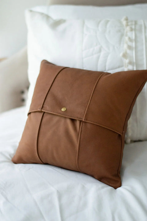 The Urban Southern Small Leather Pillow, crafted from full-grain leather with a buttoned envelope-style flap, rests on a white bed accented by crisp white quilted pillows in the background.
