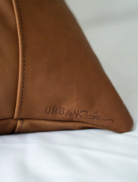 Close-up of a brown Small Leather Pillow made from full-grain leather, embossed with the Urban Southern brand name. The out-of-focus white fabric background highlights the craftsmanship of this American-made accent piece.