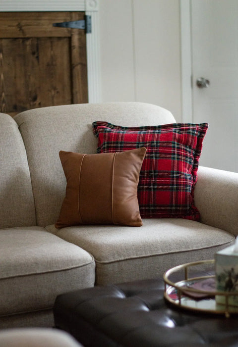 A beige sofa with an Urban Southern Small Leather Pillow and a red plaid pillow sits beside a wooden door, while a round tray with decor items rests on a dark ottoman in the foreground.