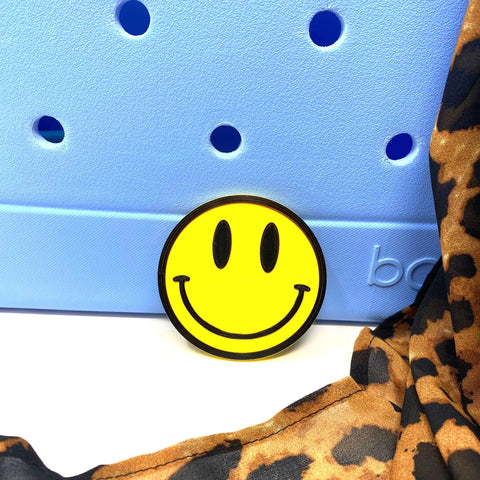 A FRESHe BOGLETS Smiley Emoji Charm by Chatelet Manufacturing is propped against a light blue perforated surface, with a brown and black leopard print fabric draped nearby. Compatible with Bogg Bags, Simply Southern, and other tote bags.