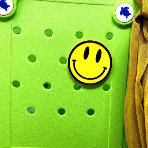 A FRESHe BOGLETS Smiley Emoji Charm by Chatelet Manufacturing is attached to a light green tote bag with circular holes, alongside two white blue-turtle buttons and yellow fabric—showcasing a fun accessory idea for Bogg Bags and more.