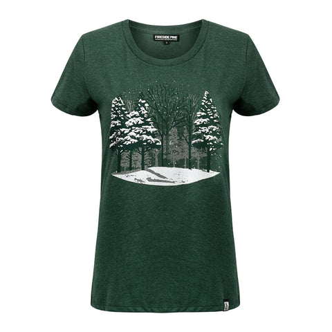 The Fireside Pine Snow Pine t-shirt is a dark green, short-sleeve shirt made in the USA, featuring a snowy winter forest scene with snow-covered trees on the front.