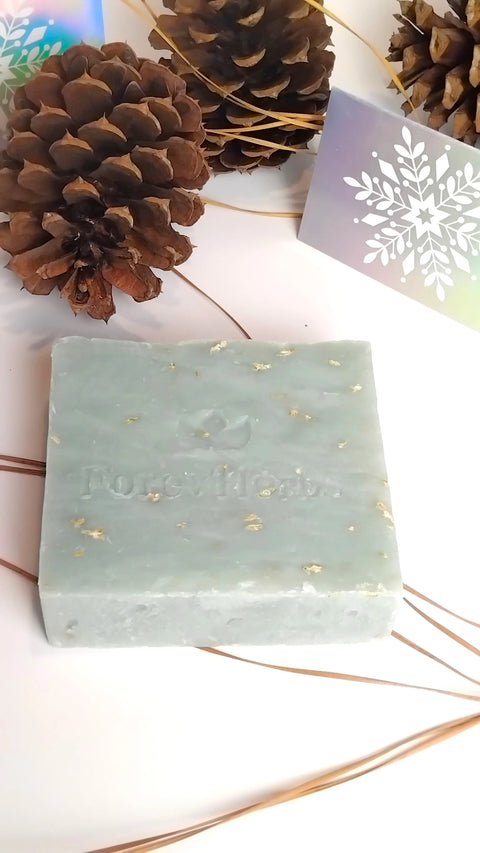 The Forev'Herbs Snowflakes Handmade Soap is a square, light green bar with gold flecks, displayed on a white surface among pinecones and winter cards—ideal for adding a festive touch to your winter skincare routine.