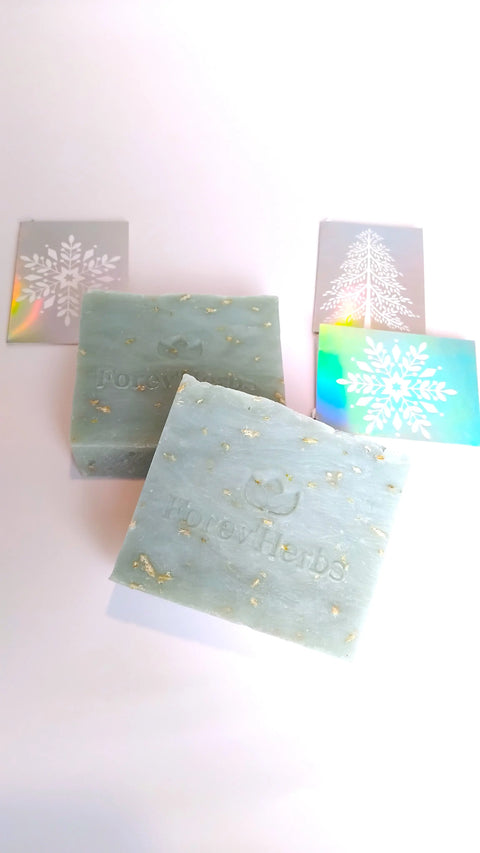 Two greenish bars of Forev'Herbs Snowflakes Handmade Soap are displayed on a white background, with two silver snowflake designs and a silver Christmas tree behind them—perfect for your winter skincare routine.