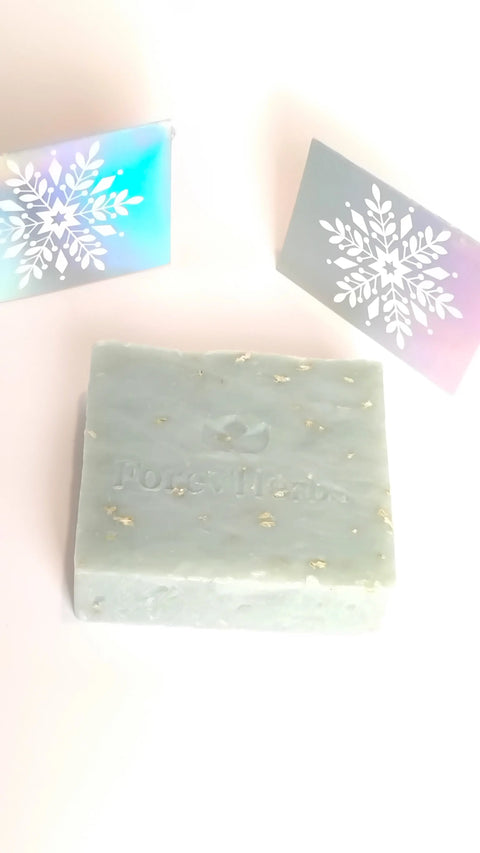 A light blue Forev'Herbs Snowflakes Handmade Soap bar with gold flecks and embossed branding, ideal for winter skincare, sits in front of two pastel boxes featuring white snowflake designs.
