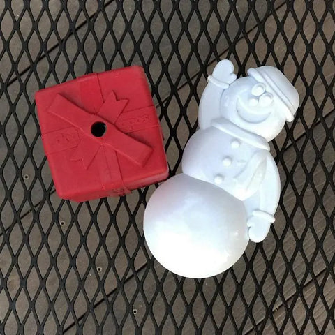 A red Gift Box EDispenser Durable Rubber Chew Toy & Treat Dispenser by SodaPup sits next to a white plastic snowman with a hat and raised hand, both on a black metal mesh surface.