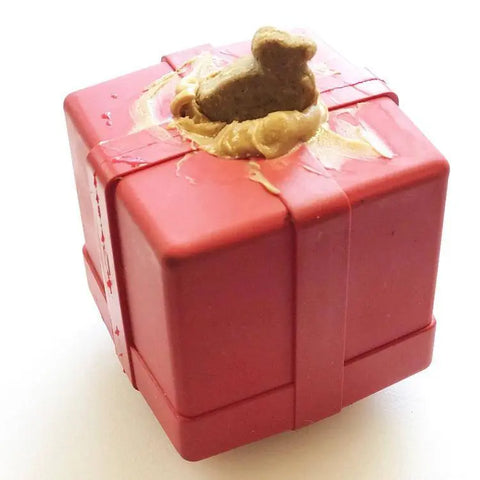 The SodaPup Gift Box EDispenser is a durable red rubber chew toy and treat dispenser, cube-shaped with peanut butter and a small bone dog treat on top—perfect as a gift for your pup.