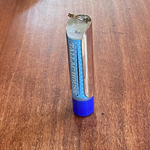 The Tallac House Lip Balm Solid Metal Keychain Holder with dial protection stands upright on a wooden surface, featuring a blue cap, silver body, and metal ring—perfect for keeping lip balm handy. Made in USA.