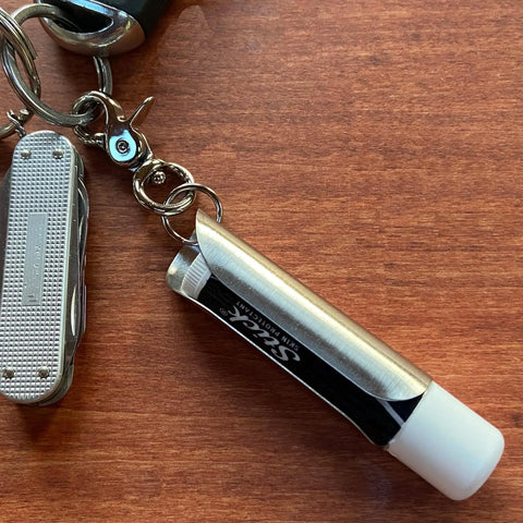 A Tallac House Lip Balm Solid Metal Keychain Keyring Holder with dial protection for lip balm, made in USA, is attached to a keyring and rests on a wooden surface.