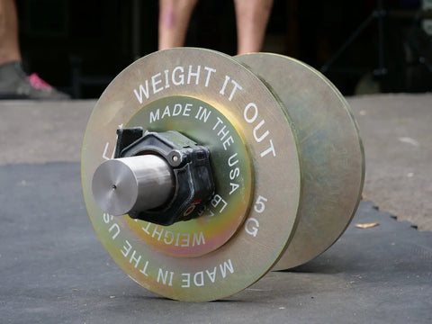 A close-up of a Solid Steel Loadable Dumbbell Set by Weight It Out, engraved with “MADE IN THE USA 5 KG,” rests on a gym floor, with blurred legs and shoes visible in the background.