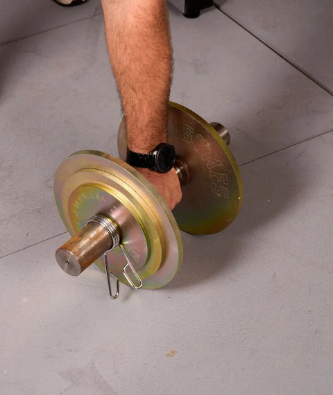 A person’s arm, wearing a black watch, grips the handle of a Weight It Out Solid Steel Loadable Dumbbell Set with calibrated plates, resting on a light gray floor.