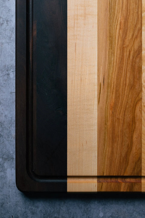 Two Montana Block The Utilitarian 14 x 20 solid wood cutting boards rest on a gray surface—one dark brown, the other an edge grain design with alternating light and medium tones. Only the corners of each are shown.