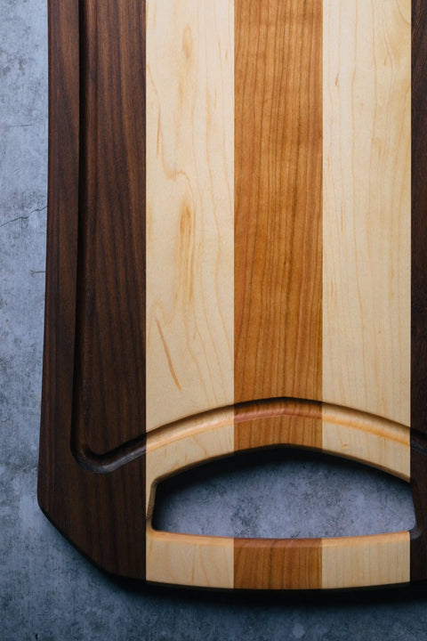 A close-up of Montana Blocks The Rectangular, a wood board with alternating dark, light, and medium stripes and a built-in handle, placed on a gray textured surface.