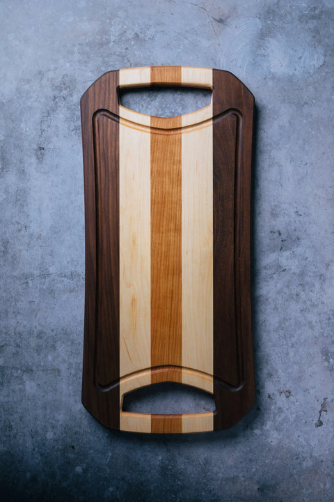 The Rectangular by Montana Block, a handmade serving tray with curved edges, two handles, and alternating light and dark wood stripes, rests on a gray textured surface.