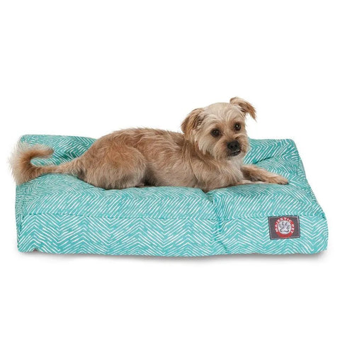 A small, light brown dog relaxes on the Majestic Pet South West Rectangle Dog Bed, featuring a turquoise and white zigzag pattern. The bed is washable for easy cleaning and sports a label with a paw print.
