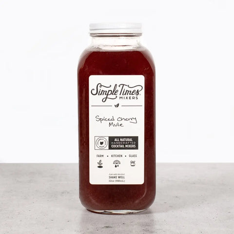 A clear glass bottle of Simple Times Mixers Spiced Cherry Mule non-alcoholic cocktail mixer with a white label sits on a gray surface against a plain white background.