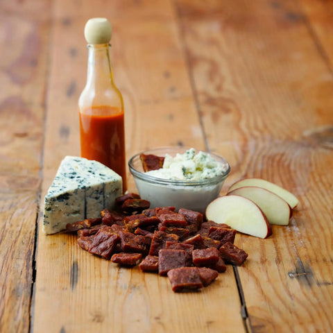 A wooden table displays sliced apples, blue cheese, hot sauce, dip, Field Trip Buffalo Style Chicken Bites 2.2oz (4ct), and small pieces of beef jerky arranged together.