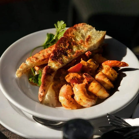 A white plate with shrimp in yellow sauce, garnished with herbs and vendor-unknown's Spicy Salts for Spring - 3 Pack Collection, is served with toasted bread. Sunlight enhances the dish's warmth and appeal.