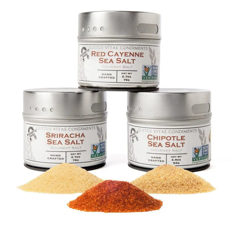 Three tins from Gustus Vitae’s Red Hot Sea Salts Collection are stacked in a pyramid, with vibrant colored salts poured in front of each tin against a white background.