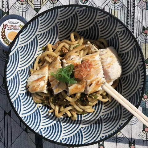 A patterned bowl of udon noodles with grilled chicken, green onions, and bonito flakes is served with chopsticks. Next to it is the Gustus Vitae Red Hot Sea Salts Collection - 3 Tins on a geometric-patterned tablecloth.